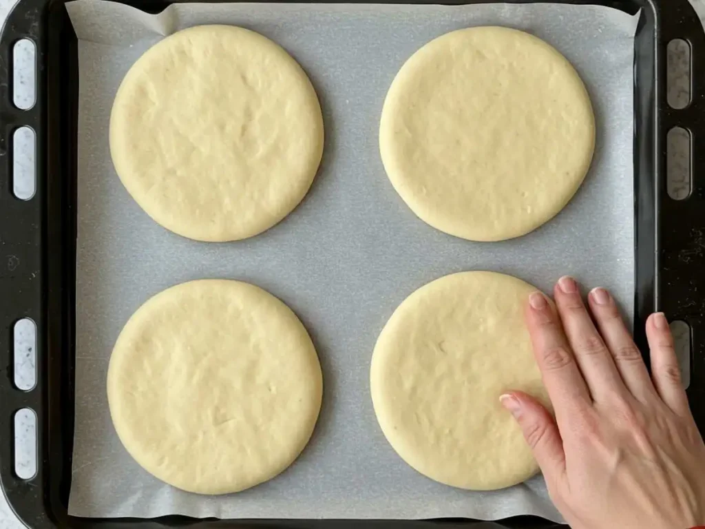 Cuatro piezas de pan piedra formadas y aplanadas sobre una bandeja de hornear, listas para la segunda fermentación.